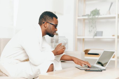 Photo for African American Man in Bathrobe Typing on Laptop while Working from Home on the Sofa He is Smiling and Enjoying a Productive Morning in a Modern Living Room The Image Represents Education, - Royalty Free Image