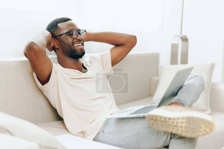 Foto de Sonriente hombre afroamericano escribiendo en el ordenador portátil mientras trabaja desde casa en un sofá moderno Un joven y feliz freelancer negro, con ropa casual, se sienta cómodamente en un sofá en su moderna sala de estar - Imagen libre de derechos