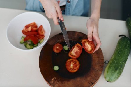 Foto de Ensalada de tomate fresca y saludable: manos de una mujer cortando verduras verdes frescas en una tabla de picar de madera con un tomate rojo. - Imagen libre de derechos