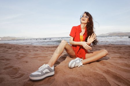 Téléchargez les photos : Femme souriante profitant de vacances à la plage insouciantes, assise sur le sable, avec la vue grand angle sur la belle toile de fond de mer d'été. - en image libre de droit