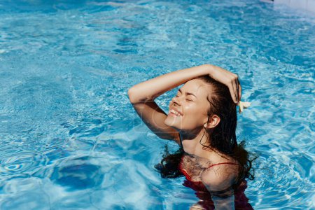 Foto de Mujer joven en la piscina en traje de baño rojo con hermosa. sonrisa al sol corrige su cabello con la mano, la belleza y el bronceado. Foto de alta calidad - Imagen libre de derechos