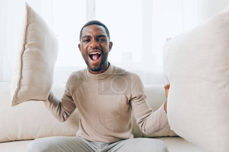 Téléchargez les photos : Homme afro-américain détendu assis sur un canapé confortable dans son salon moderne, tenant un oreiller et souriant Il profite d'un week-end paisible à la maison, pensant et regardant quelque chose en ligne - en image libre de droit