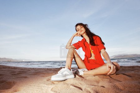 Téléchargez les photos : Mode plage : Femme souriante assise sur le sable, embrassant les vibrations estivales avec une tenue à la mode et un style de vie sans soucis - en image libre de droit