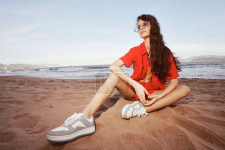 Téléchargez les photos : Femme souriante en tenue tendance, assise sur le sable à la plage, profitant de vacances insouciantes : Beachside Happiness - en image libre de droit
