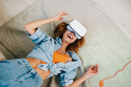 Foto de Mujer sonriente disfrutando del juego de realidad virtual en casa con gafas VR futuristas y camisa blanca, sentada en un sofá en una sala de estar moderna y digital. - Imagen libre de derechos