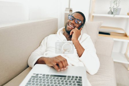 Foto de Sonriente hombre afroamericano trabajando en un ordenador portátil en una acogedora sala de estar La imagen muestra a un freelancer alegre y enfocado, con un albornoz, sentado en un sofá con un ordenador portátil en su regazo - Imagen libre de derechos