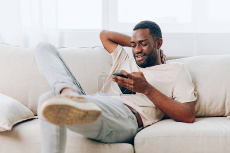 Foto de Feliz hombre afroamericano sentado en un sofá negro moderno, relajándose en casa y utilizando su teléfono inteligente Está escribiendo un mensaje, totalmente inmerso en el mundo de la tecnología y la comunicación El acogedor - Imagen libre de derechos