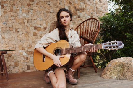 Photo for A young woman sitting on a wooden deck with an acoustic guitar in front of a stone wall - Royalty Free Image