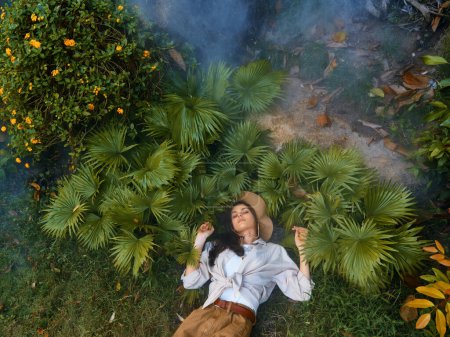 Téléchargez les photos : Une femme dans un chapeau étendu dans l'herbe à côté d'un palmier avec de la fumée qui en sort - en image libre de droit