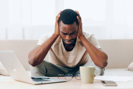 Téléchargez les photos : Fatigué afro-américain pigiste travaillant sur ordinateur portable dans le Home Office, se sentant malheureux et déprimé sur le canapé - en image libre de droit