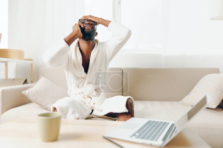 Foto de Freelancer afroamericano sonriente que trabaja en el ordenador portátil en la acogedora sala de estar con computadora portátil y teléfono en el sofá - Imagen libre de derechos
