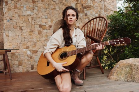 Téléchargez les photos : Une jeune femme assise sur une terrasse en bois avec une guitare acoustique devant elle - en image libre de droit