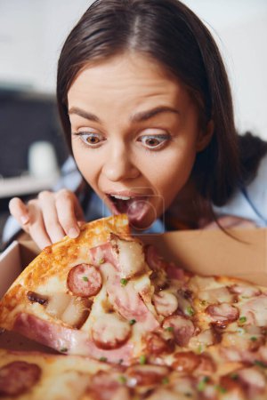Photo for Hungry woman holding pizza box with mouth wide open, eagerly reaching for a slice of pizza in front of her - Royalty Free Image