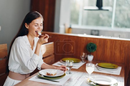 Foto de Mujer sonriente disfrutando de una cena romántica en casa Hermosa morena sentada en la mesa de comedor de moda, deleitándose con pastel y vino caseros - Imagen libre de derechos
