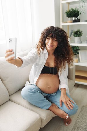 Téléchargez les photos : Femme enceinte blogueuse assise sur le canapé à la maison et prend des photos d'elle-même au téléphone, selfie et appel vidéo, consultation avec le médecin en ligne, la gestion de la grossesse. Photo de haute qualité - en image libre de droit