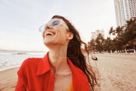 Téléchargez les photos : Femme souriante embrassant la liberté : portrait coloré d'un modèle à la mode avec bouche ouverte et lunettes de soleil, profiter de vacances sans soucis à la plage du coucher du soleil - en image libre de droit