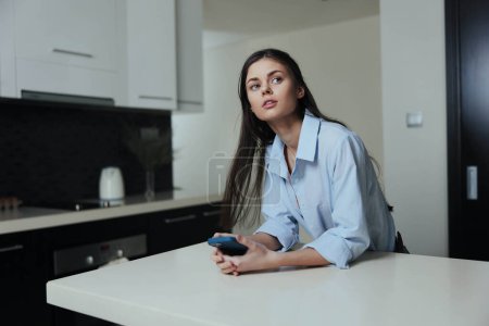 Téléchargez les photos : Femme assise sur le comptoir dans la cuisine avec téléphone portable à la main, profitant d'un moment de détente - en image libre de droit