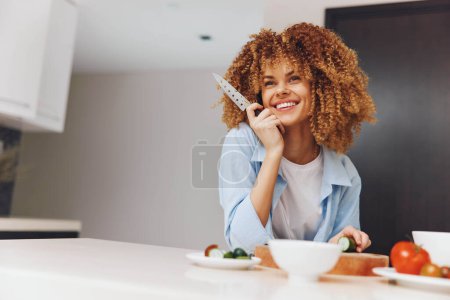 Téléchargez les photos : Heureuse femme afro-américaine utilisant un téléphone portable à la table de cuisine, souriante et engagée dans une conversation - en image libre de droit