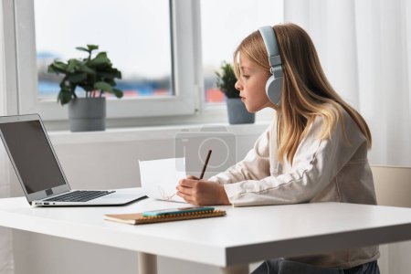 Photo for Smiling girl learning at home with a laptop elearning in action The cute, young Caucasian schoolgirl sits at a desk, immersed in her online education With headphones on, she engages in a virtual - Royalty Free Image