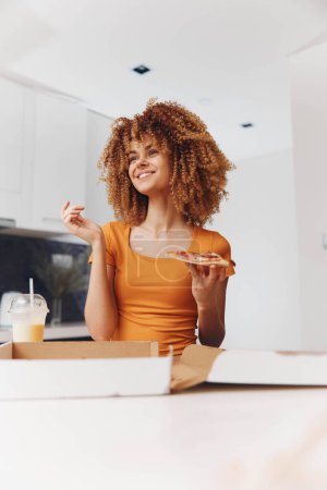 Photo for Attractive woman with curly hair enjoying a slice of pizza in her hand while wearing an orange shirt - Royalty Free Image