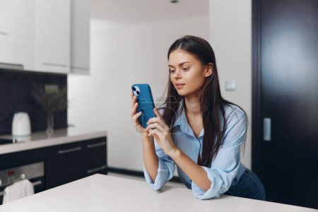 Photo for Young woman using smartphone while sitting on kitchen counter at home - Royalty Free Image