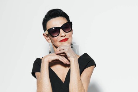 Photo for Fashionable woman in black dress and sunglasses posing for the camera with hand on chin - Royalty Free Image