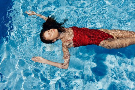 Téléchargez les photos : Jeune femme dans la piscine en maillot de bain rouge avec un beau sourire couché sur l'eau et nager au soleil dans la piscine, le concept de détente en vacances. Photo de haute qualité - en image libre de droit