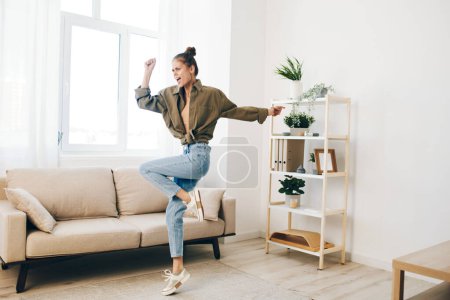 Foto de Mujer alegre saltando y bailando en un apartamento casero, disfrutando de la música y la relajación en el sofá" - Imagen libre de derechos