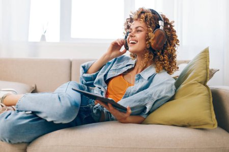 Foto de Happy Woman Holding Tablet PC en casa, lectura, escuchar música con auriculares, sentarse en el sofá en la acogedora sala de estar. - Imagen libre de derechos