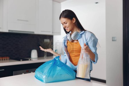 Téléchargez les photos : Jeune femme tenant un sac de déchets devant le comptoir de cuisine, concept de recyclage et d'élimination des déchets - en image libre de droit