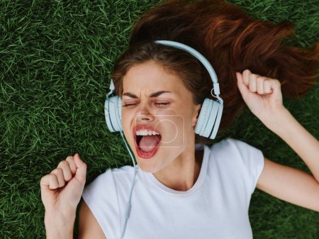Téléchargez les photos : Belle femme dans les écouteurs écouter de la musique couché sur la pelouse verte de l'herbe dans le parc en été et rire, bonheur dans la nature, mode de vie d'été. Photo de haute qualité - en image libre de droit