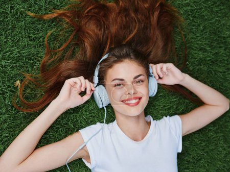Téléchargez les photos : Joyeux adolescent écoutant de la musique avec des écouteurs dans un T-shirt blanc sur l'herbe verte de la pelouse dans le parc, sourire de vacances avec des dents, regardant dans la caméra. Photo de haute qualité - en image libre de droit