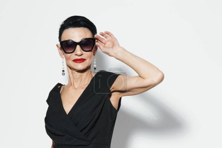 Téléchargez les photos : Elégante femme âgée en robe noire et lunettes de soleil posant pour caméra devant un mur blanc - en image libre de droit