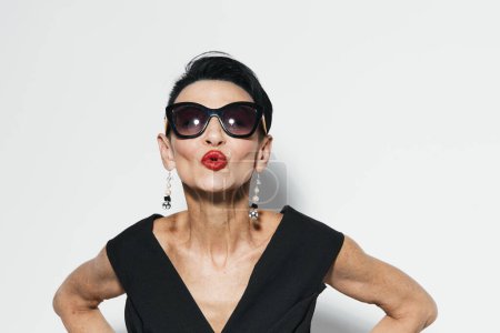Téléchargez les photos : Femme sexy dans des lunettes de soleil élégantes et robe noire frappant une pose drôle avec les mains sur les hanches - en image libre de droit
