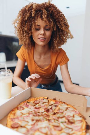 Foto de Feliz joven afroamericana mujer disfrutando deliciosa pizza para el almuerzo en su acogedora cocina casera - Imagen libre de derechos