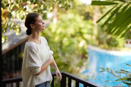 Foto de Mujer disfrutando de unas vacaciones relajantes en un balcón tropical, sonriendo bajo la cálida luz del sol Ella se encuentra junto a la piscina, rodeada de plantas exuberantes, saboreando una bebida refrescante y la libertad de - Imagen libre de derechos