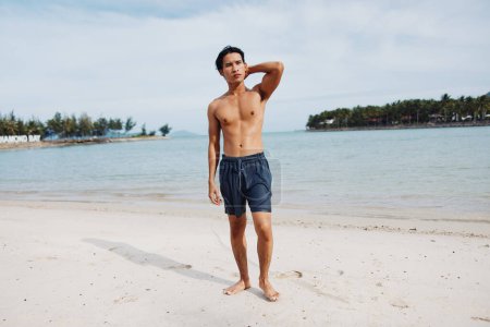 Photo for Smiling Asian Man Enjoying Beach Vacation: Muscular Torso and Happy Smile against Tropical Island Background - Royalty Free Image