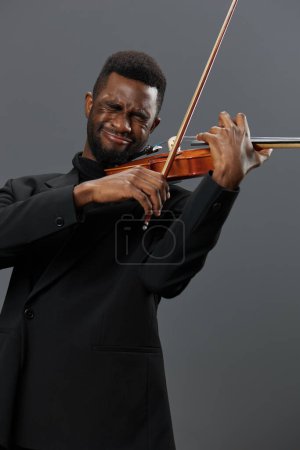 Foto de Elegante hombre afroamericano en esmoquin tocando el violín sobre un fondo gris, creando música hermosa - Imagen libre de derechos