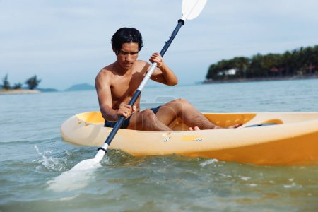 Foto de Kayak Adventure: Un feliz asiático disfrutando de vacaciones de verano en una playa tropical, remando con un remo en su canoa - Imagen libre de derechos