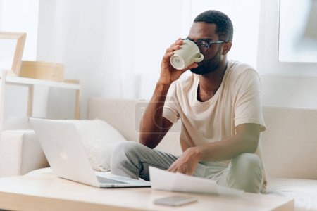 Téléchargez les photos : Homme afro-américain souriant travaillant sur ordinateur portable dans le bureau moderne, dactylographier et profiter du style de vie freelance Le jeune homme noir est assis sur un canapé confortable dans son salon appartements, entouré - en image libre de droit