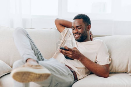 Téléchargez les photos : Afro-Américain assis sur un canapé noir, heureux d'utiliser son smartphone pour discuter et se connecter avec des amis via les médias sociaux Il est détendu et confiant, profiter de son temps libre à la maison Le moderne - en image libre de droit