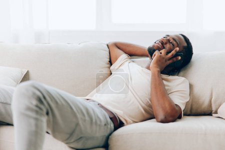 Téléchargez les photos : Heureux Afro-Américain assis sur un canapé noir, tenant un téléphone portable et profitant d'un appel vidéo sur son canapé moderne dans le confort de sa maison - en image libre de droit
