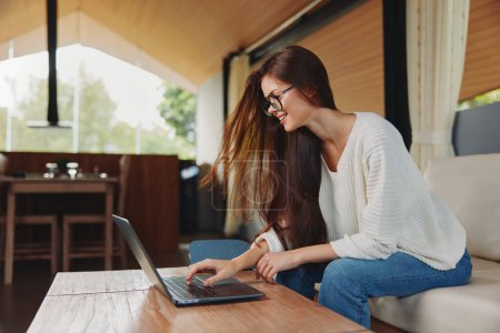 Photo for Relaxed Millennial Woman Sitting on a Sofa with Laptop at Home Smiling and Teaching Online She is wearing glasses and surrounded by a cozy living room Her notebook and computer show that she is - Royalty Free Image