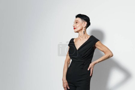 Téléchargez les photos : Femme mûre élégante en robe noire élégante posant avec confiance avec les mains sur les hanches devant le mur blanc - en image libre de droit