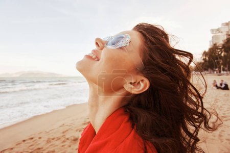 Téléchargez les photos : Femme souriante à la plage, profiter de la brise de la mer et le coucher du soleil Faire place à la liberté et le bonheur dans les lunettes de soleil à la mode - en image libre de droit