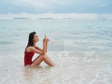 Photo for Woman with a beautiful tan tourist in a red swimsuit sitting on the sand on the beach in the ocean in the waves pensive. High quality photo - Royalty Free Image