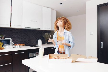 Téléchargez les photos : Femme debout dans la cuisine avec une pizza et une boîte de beignets devant elle, préparant de délicieuses friandises maison - en image libre de droit