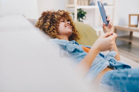 Foto de Mujer feliz sentada en un sofá acogedor, sosteniendo el teléfono móvil y sonriendo mientras disfruta de juegos en línea y leyendo mensajes en una hermosa casa. - Imagen libre de derechos
