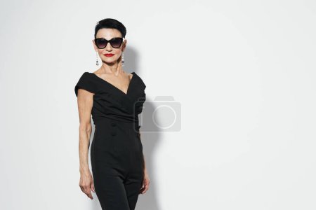Photo for Stylish woman in black dress and sunglasses posing confidently in front of white wall - Royalty Free Image