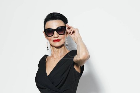 Foto de Mujer de edad avanzada con estilo en vestido negro y gafas de sol posando para la cámara con la mano en la oreja, retrato de señora madura de moda - Imagen libre de derechos